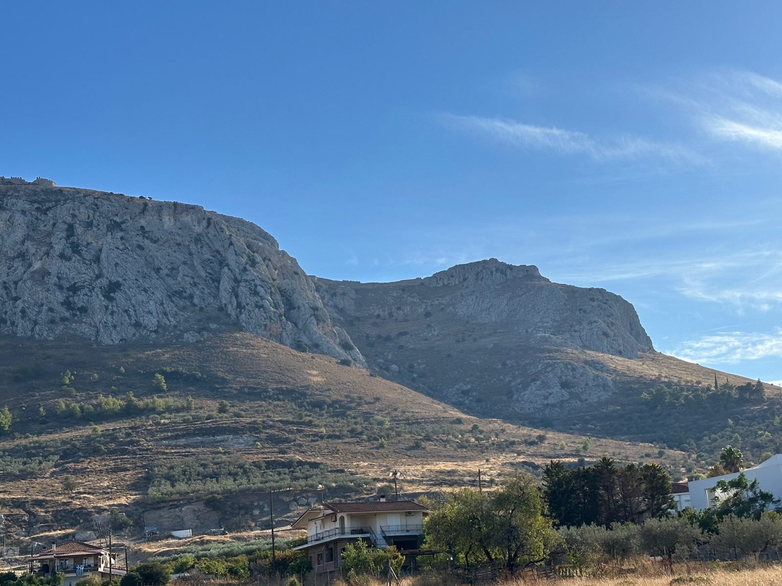 Acrocorinth, the fortress rock above Corinth, with olive trees and dry hillside below. Walls run along the summit; Greeks, Romans, Byzantines, Franks, Venetians, and Turks all held it in turn, because whoever held this rock held the road between northern Greece and the Peloponnese. The soil is thin and sun-baked, and a handful of defenders on a height like this could hold the only road against an army. Rock like this split Greece into hundreds of separate city-states.