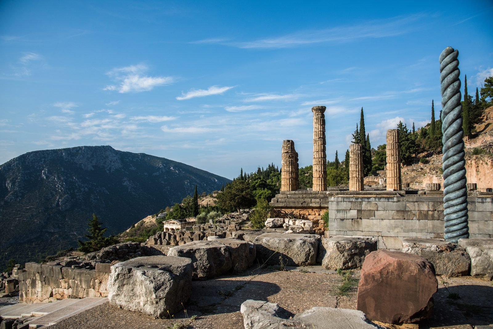 The Serpent Column at Delphi, with the columns of the Temple of Apollo behind it and the mountains across the valley. Three bronze serpents twist together into one column, cast from the Persian weapons captured at the battle of Plataea in 479 BC and inscribed with the names of the thirty-one Greek cities that fought there together. This is a modern copy standing where the original stood; the emperor Constantine carried the original off to Constantinople, where it still stands. Thirty-one rival cities, one column, one list of names: that is kleos, glory written down so it cannot be erased, as the name of Koroibos was.