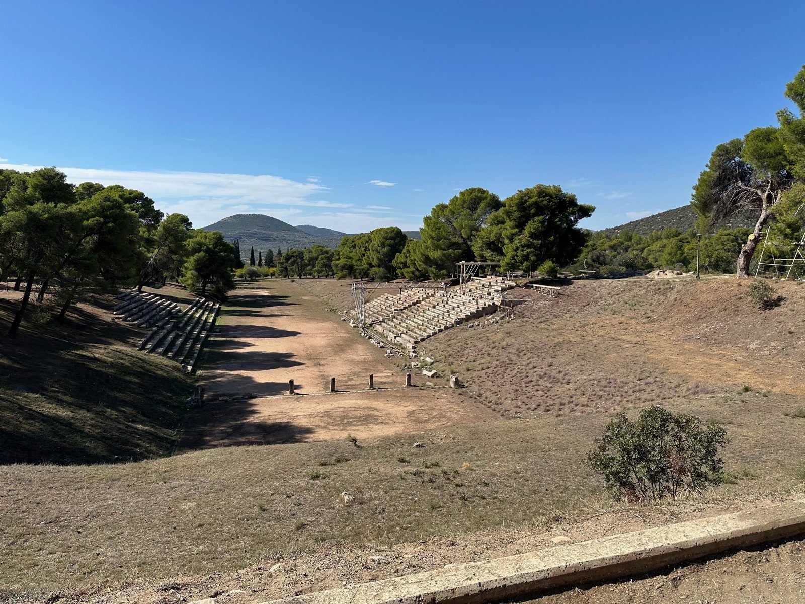 The stadium at Epidaurus in the Peloponnese, with mountains closing it in on every side. The low row of stones across the track in the foreground is a starting line, the balbis, with grooves cut for a runner's bare toes. This is not Olympia, and this track was laid out about 400 BC, nearly four centuries after Koroibos ran. It is here because I have stood my students on a line like this one, in a valley like this one, and asked the question this lesson asks: what made a thousand warring cities one people? A race, it turns out, had more to do with the answer than any king.